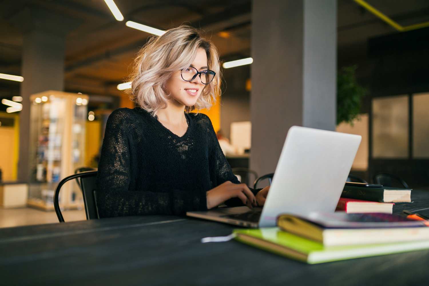 portrait-of-young-pretty-smiling-woman-sitting-at-table-in-black-shirt-working-on-laptop-in-co-working-office-wearing-glasses